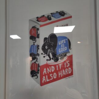 Photo with reflection from lights of print by Anna May Henry of a box that appears like the candy coated peanuts candy with an elephant in the middle, a blanket over the elephant reads Life is Good and below reads And It Is Also Hard